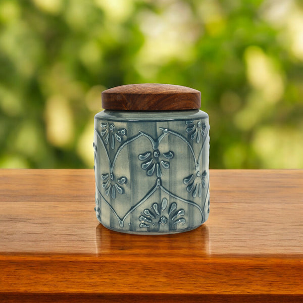 A blue ceramic air-tight jar with intricate flower embossed etchings, featuring a wooden lid, placed on a wooden surface.