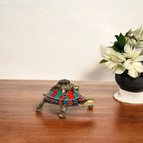 A brass tortoise figurine with red and blue patterns on its shell, positioned on a wooden surface with a vase of white flowers in the background.