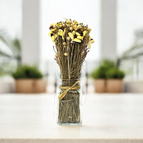A handcrafted Sholapith dried flower bouquet in a glass jar, with soft color tulip buds and dried grass stems, tied with rustic raffia.