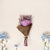 A mini dried flower bouquet with a lavender rose, wrapped in brown kraft paper with natural twine, surrounded by decorative blue and white flowers.