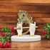 A polyresin statue of Krishna's hands holding a flute, with gold detailing on the statue, placed on a white base, surrounded by red and green foliage.