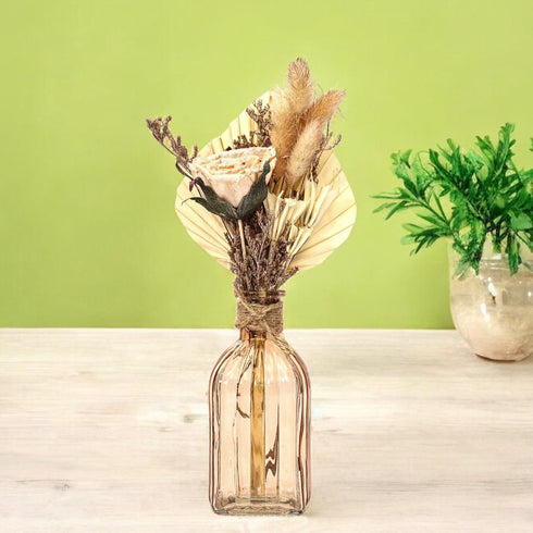 A rustic dried flower bouquet arranged in an amber glass bottle, featuring pampas grass, palm fan leaves, and a preserved rose, tied with natural jute rope.