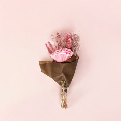 A small dried flower bouquet featuring pink roses, wrapped in brown kraft paper with a twine tie on a pink background.