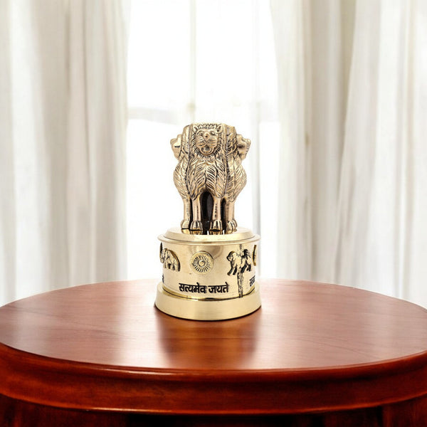 An antique brass Ashok Stambh showpiece, featuring four lions and the Dharma Chakra, placed on a circular base with inscriptions, displayed on a wooden surface.