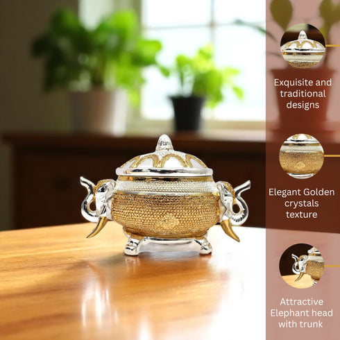 Decorative golden and silver container with elephant heads on a wooden surface, surrounded by plants.