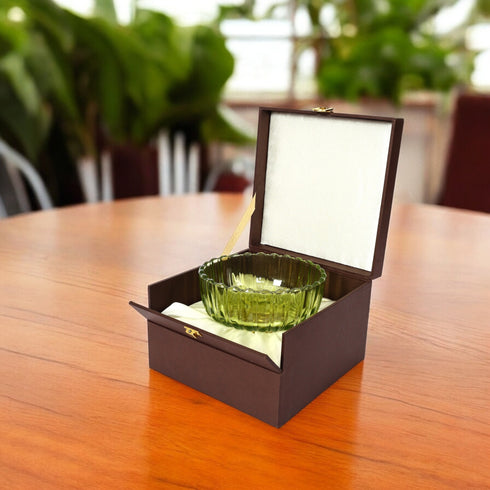 Green glass bowl in an open brown box on a wooden table with blurred greenery in the background