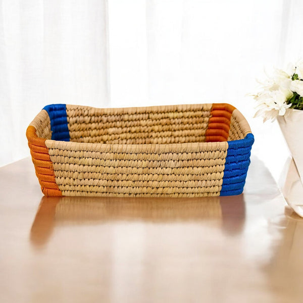 A handwoven Sabai Grass storage organizer with beige body and blue and orange trim, placed on a table with a floral arrangement in the background.