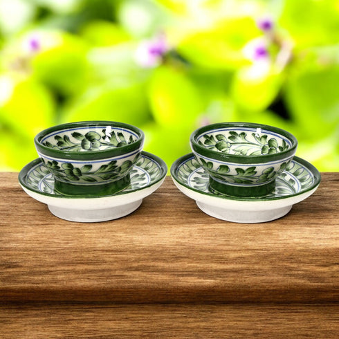 Handcrafted Blue Pottery bowl and plate set with floral motifs on a green and white background, placed on a wooden surface with foliage in the background.