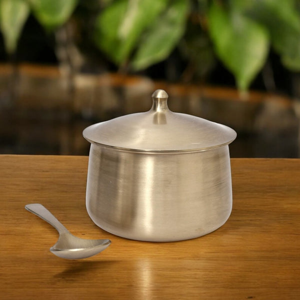 Stainless steel pot with lid and spoon on a wooden surface with a blurred green plant background