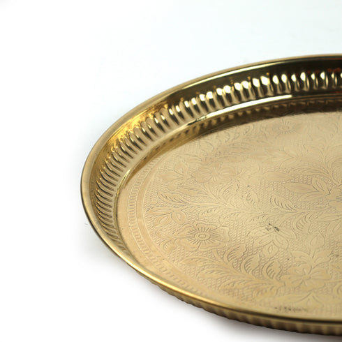 A close-up of a round brass tray with a fluted rim and an engraved floral pattern on the surface, set against a plain white background.