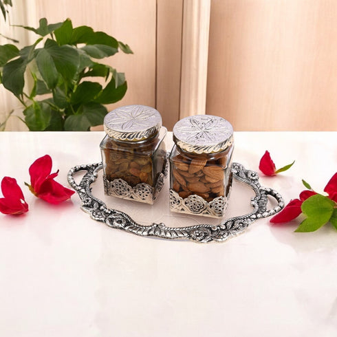 Two decorative glass jars with silver lids on a white surface with red flowers and green leaves.