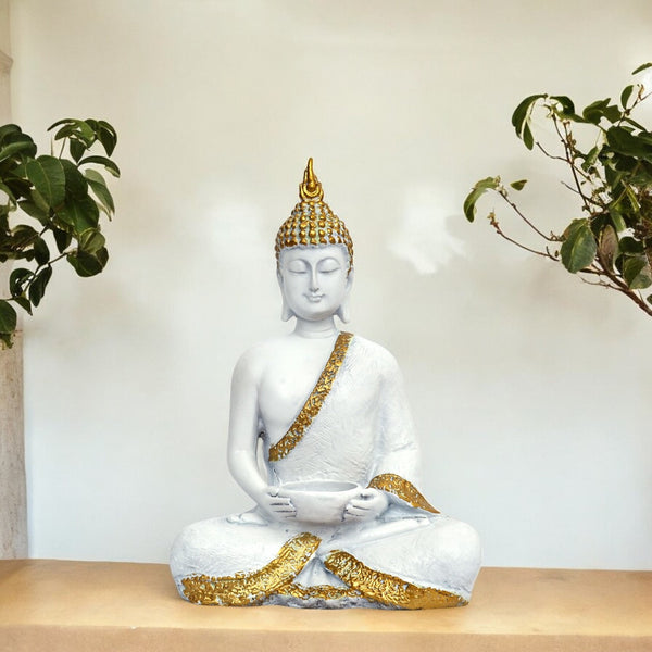 White Buddha statue with gold accents on a wooden surface with plants in the background