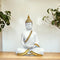 White Buddha statue with gold accents on a wooden surface with plants in the background