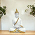 White Buddha statue with gold accents on a wooden surface with plants in the background