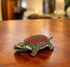 A brass tortoise figurine with a decorative red and green pattern on its back, displayed on a wooden surface.