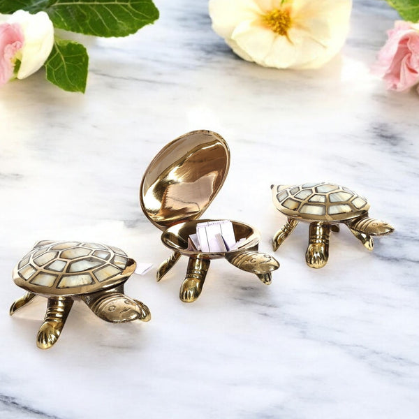 A set of three brass tortoise figurines with a secret wish compartment, placed on a marble surface with flowers in the background.