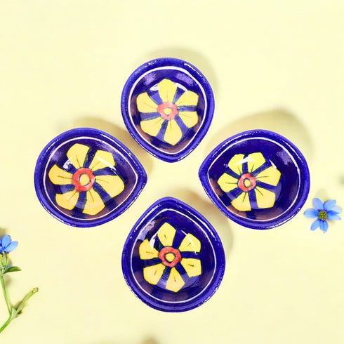 A set of four handcrafted blue pottery diya holders with bright yellow floral designs on a deep blue base, displayed on a neutral background with a few scattered flower petals.