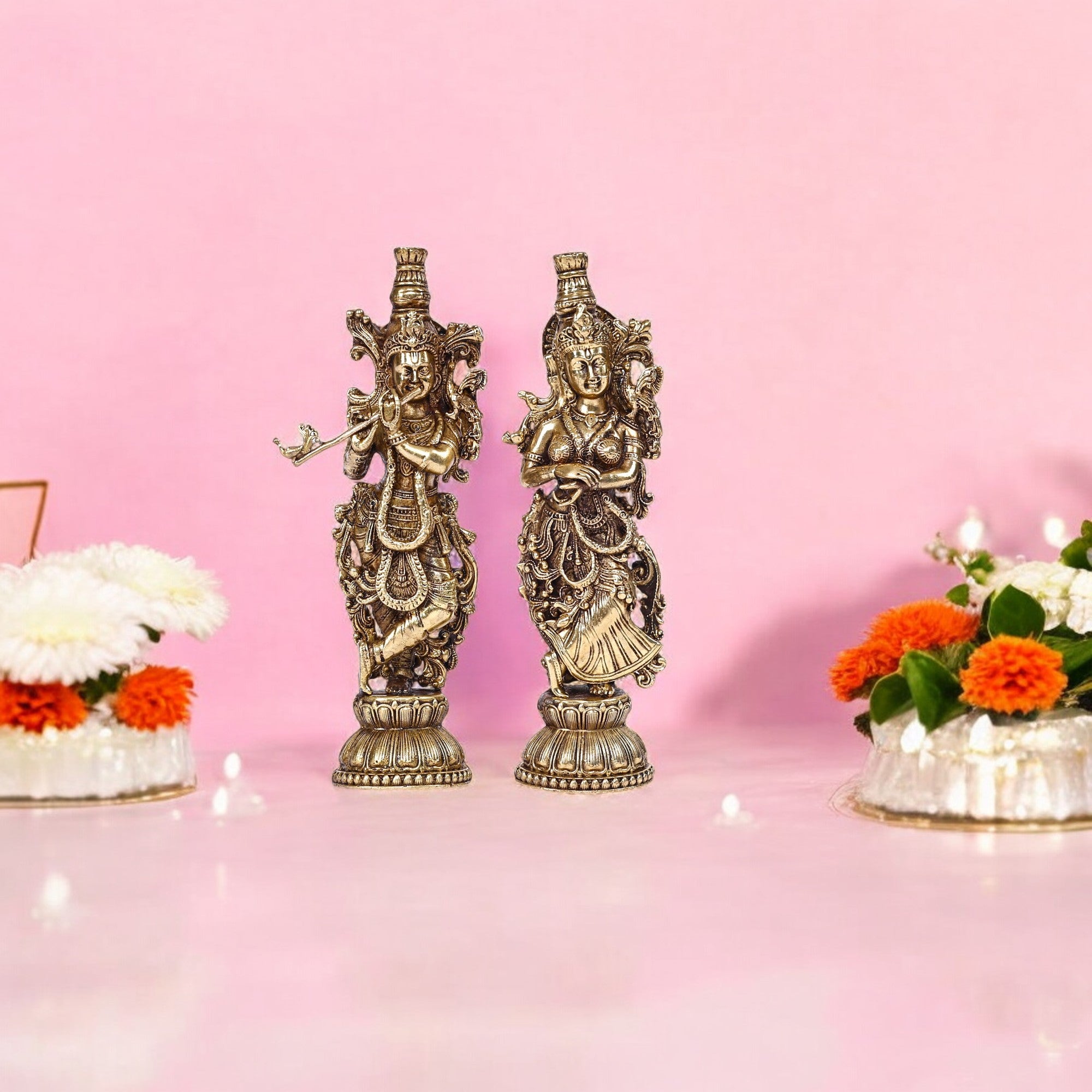 A pair of brass figurines depicting Radha and Krishna with intricate carvings, displayed on a platform with floral offerings in the background.