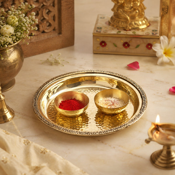 A round brass ritual tray with engraved edges and two small bowls on top, one filled with red powder and the other with a light powder, styled on a marble surface with decorative flowers, a brass pot, and a small lit diya.