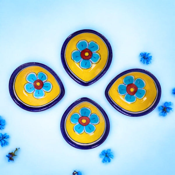 A set of four blue and yellow ceramic diyas with hand-painted floral motifs, displayed on a blue background with small blue flowers scattered around.