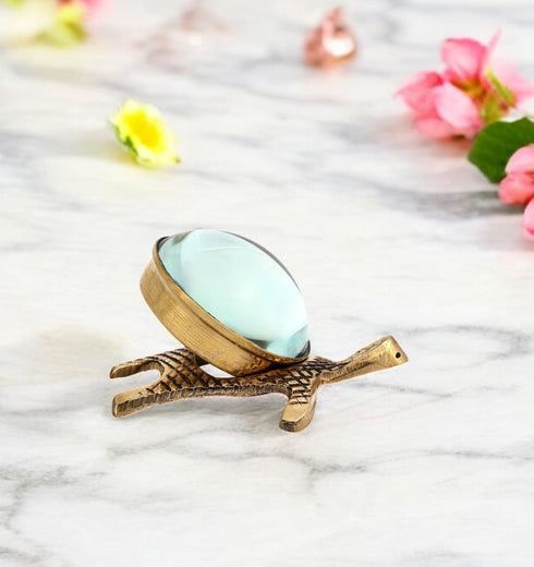A small brass tortoise-shaped Yantra with a tilted glass lens on top, placed on a marble surface surrounded by scattered flowers.