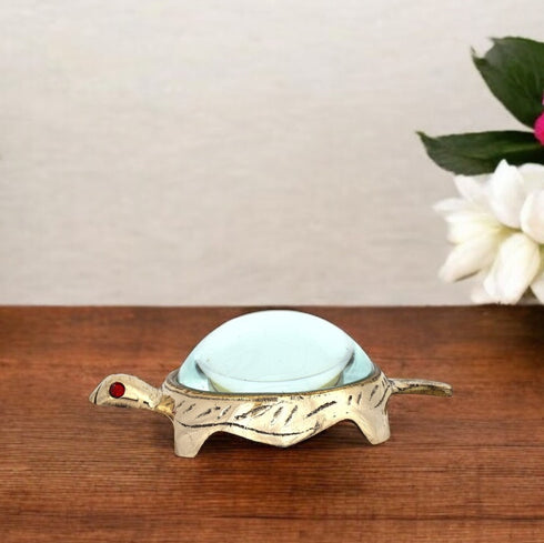A small brass tortoise-shaped yantra with a straight glass lens on a wooden surface, with a floral decoration in the background.