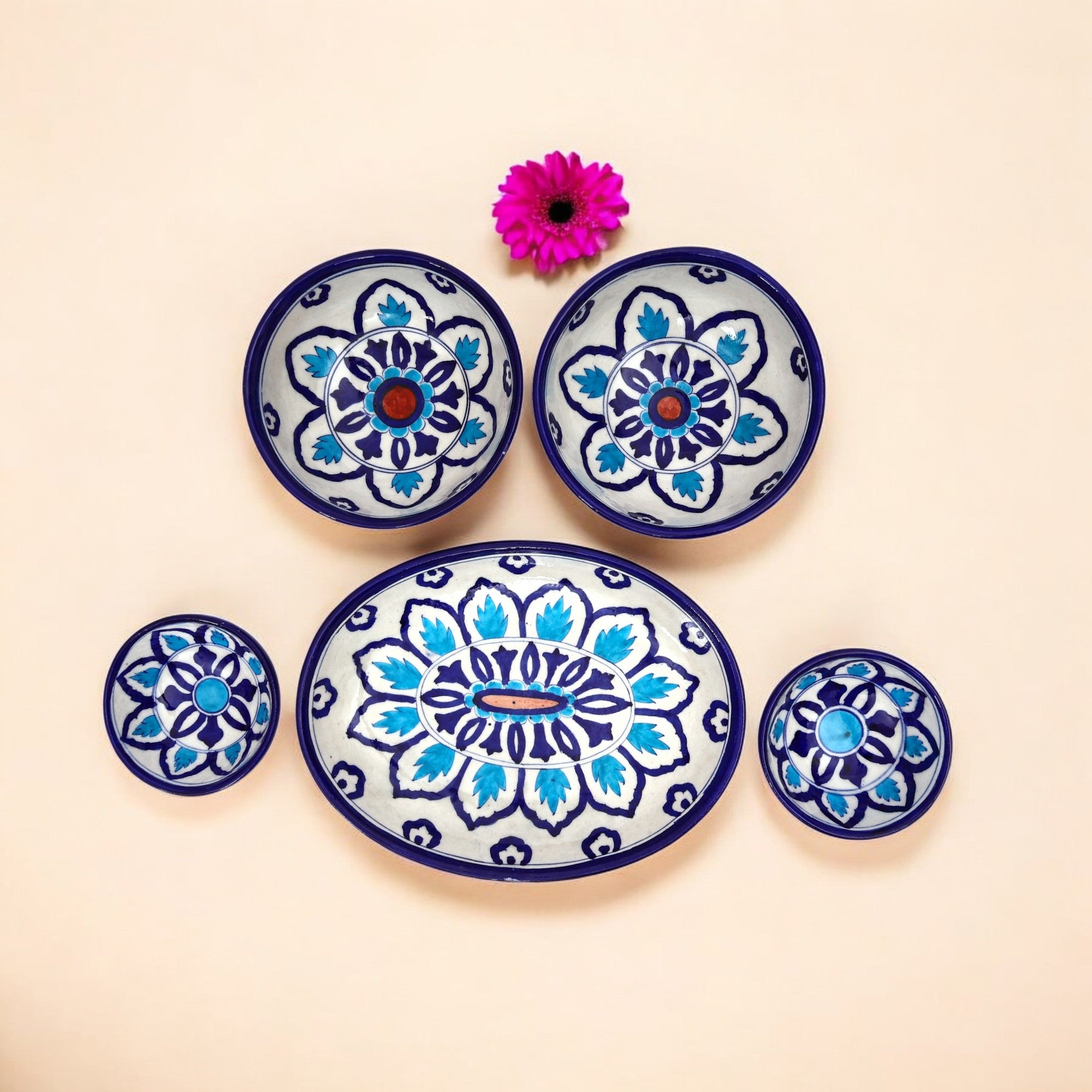 A set of handcrafted blue and white floral patterned ceramic dishes, including five pieces displayed on a neutral background with a pink flower.